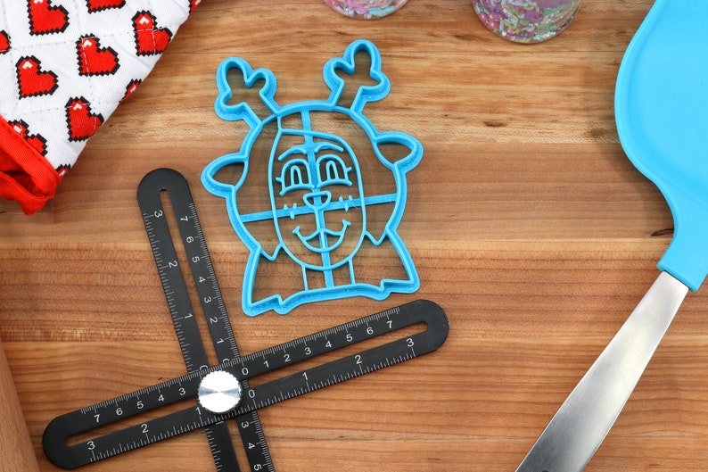 Peut inclure: Un emporte-pi&egrave;ce en plastique bleu en forme de renne avec un visage souriant et des bois. Un outil de mesure multi-angles noir est positionn&eacute; pr&egrave;s du cutter. Un gant de cuisine &agrave; motifs de c&oelig;urs rouges et blancs est visible.
