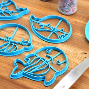 Peut inclure: Un ensemble de cinq emporte-pi&egrave;ces en plastique bleu en forme de cr&eacute;atures marines. Les emporte-pi&egrave;ces sont sur une surface en bois avec une spatule bleue.