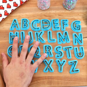 May include: A set of blue plastic alphabet cookie cutters on a wooden surface. The cutters are arranged in alphabetical order.
