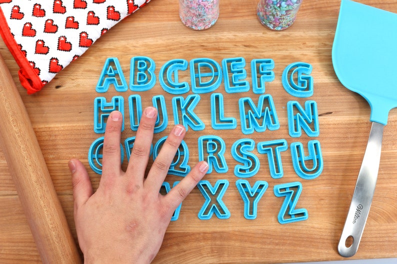 May include: A set of blue plastic alphabet cookie cutters on a wooden surface. The cutters are arranged in alphabetical order.