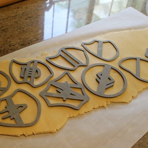 May include: A sheet of cookie dough with various metal cookie cutters pressed into it. The cutters are shaped like shields, circles, and other geometric shapes.