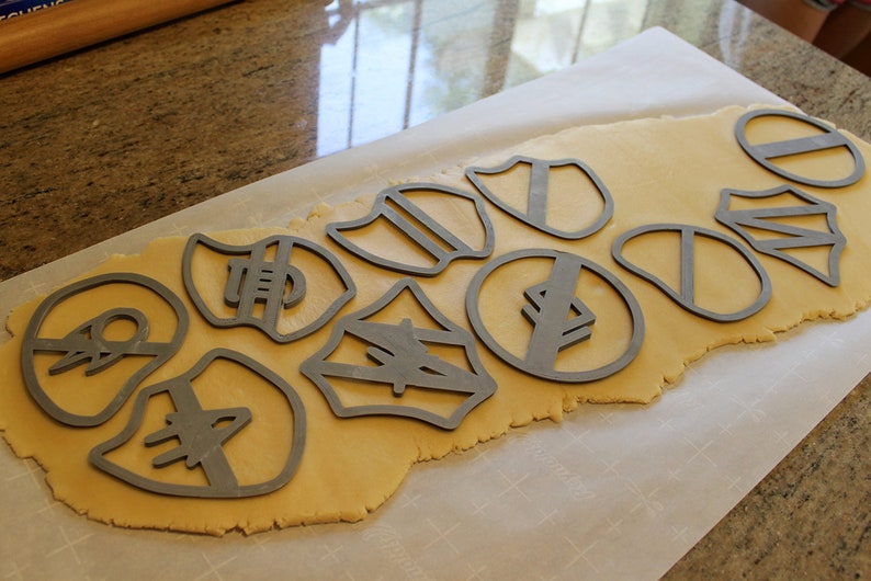 May include: A sheet of cookie dough with various metal cookie cutters pressed into it. The cutters are shaped like shields, circles, and other geometric shapes.