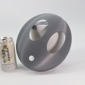 May include: A gray 3D printed object with three holes cut out. The object is shaped like a dome and is sitting on a white surface. A can of Japanese beer is sitting to the left of the object.