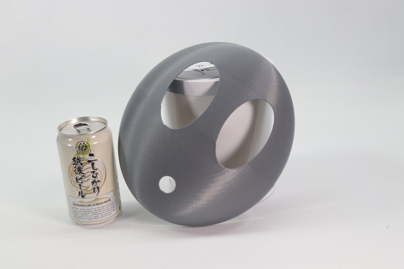 May include: A gray 3D printed object with three holes cut out. The object is shaped like a dome and is sitting on a white surface. A can of Japanese beer is sitting to the left of the object.