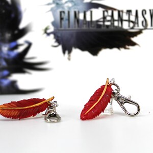 May include: Two red and gold feather charms with silver keychains. The feathers are detailed with a textured surface and a gold accent. The charms are attached to silver keychains with a lobster clasp.