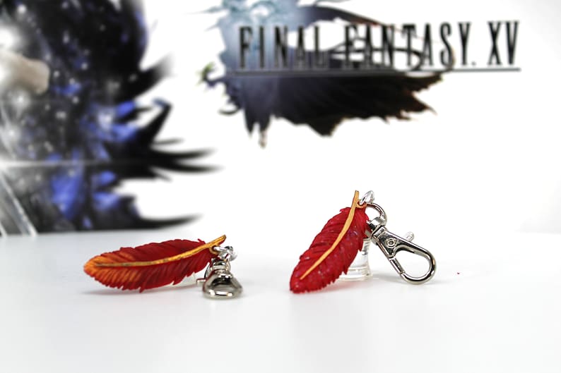 May include: Two red and gold feather charms with silver keychains. The feathers are detailed with a textured surface and a gold accent. The charms are attached to silver keychains with a lobster clasp.