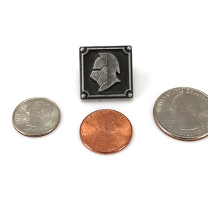 May include: A silver-colored pin with a knight's helmet design in a square frame. The pin is shown next to a US quarter and a US penny for size comparison.