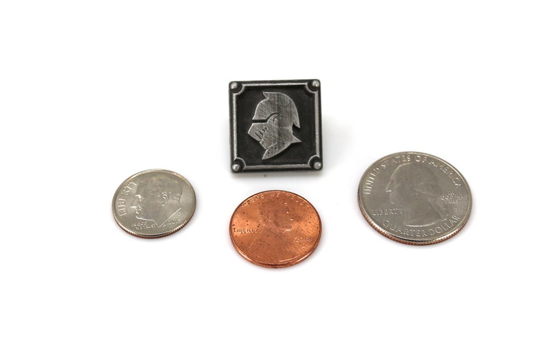 May include: A silver-colored pin with a knight's helmet design in a square frame. The pin is shown next to a US quarter and a US penny for size comparison.