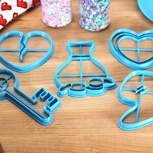 May include: Five blue plastic cookie cutters in the shapes of a key, a heart, a vase, a broken heart, and a leaf. The cutters are on a wooden surface.