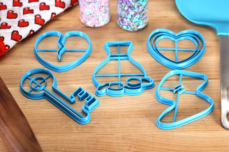 May include: Five blue plastic cookie cutters in the shapes of a key, a heart, a vase, a broken heart, and a leaf. The cutters are on a wooden surface.