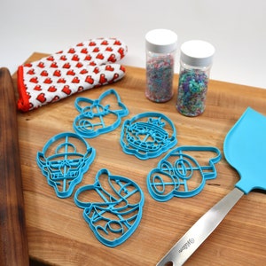 May include: A set of blue plastic cookie cutters shaped like characters from a popular video game. The cutters are on a wooden cutting board with a rolling pin, a blue spatula, and two jars of sprinkles.
