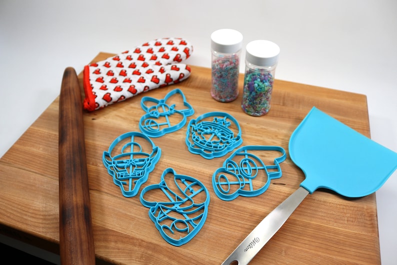 May include: A set of blue plastic cookie cutters shaped like characters from a popular video game. The cutters are on a wooden cutting board with a rolling pin, a blue spatula, and two jars of sprinkles.