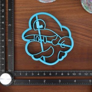 May include: Blue plastic cookie cutter shaped like a video game character with a mustache and a hat. The character has a large 'L' on the front of the hat.