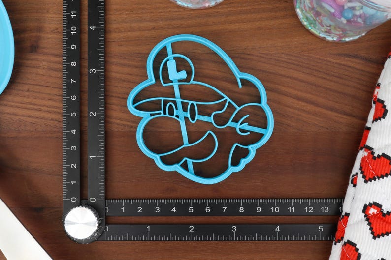 May include: Blue plastic cookie cutter shaped like a video game character with a mustache and a hat. The character has a large 'L' on the front of the hat.