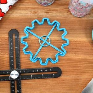 May include: A blue gear-shaped cookie cutter, a black angle ruler, and a blue spatula are on a wooden surface. A white cloth with red pixelated hearts and two jars of sprinkles are also visible. Baking and crafting supplies.