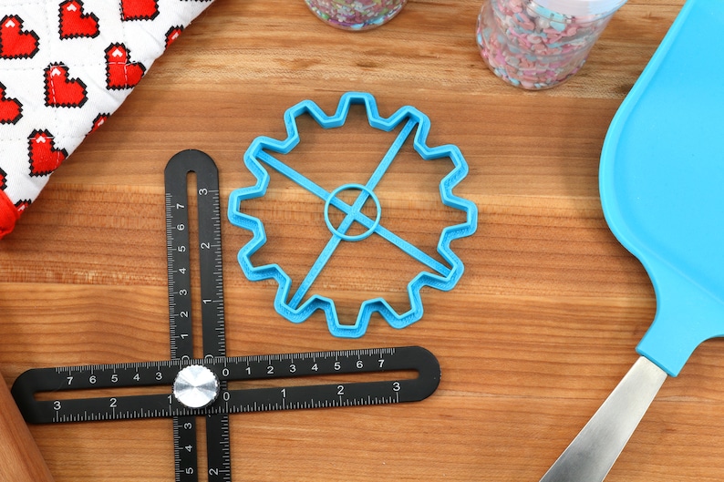 May include: A blue gear-shaped cookie cutter, a black angle ruler, and a blue spatula are on a wooden surface. A white cloth with red pixelated hearts and two jars of sprinkles are also visible. Baking and crafting supplies.
