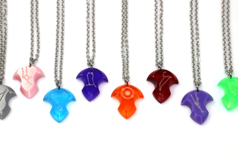 Puede incluir: Siete collares con cadenas plateadas y colgantes coloridos en forma de flecha estilizada con un dise&ntilde;o de constelaci&oacute;n. Los colgantes son de color gris, rosa, azul, morado, naranja, rojo y verde.