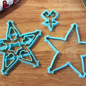 Pu&ograve; includere: Tre formine per biscotti a forma di stella azzurra su una superficie di legno. Una formina ha un intricato disegno a stella all'interno della forma a stella. Un piccolo tessuto a motivi di cuore &egrave; sullo sfondo.
