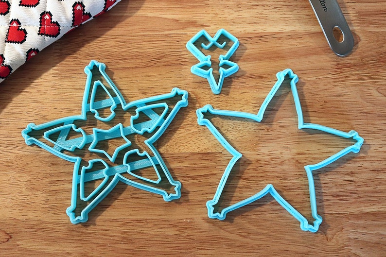 Pu&ograve; includere: Tre formine per biscotti a forma di stella azzurra su una superficie di legno. Una formina ha un intricato disegno a stella all'interno della forma a stella. Un piccolo tessuto a motivi di cuore &egrave; sullo sfondo.