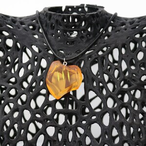 May include: A pendant necklace featuring a translucent, amber-colored stone with a unique shape. The stone hangs from a black cord and is displayed on a black, openwork mannequin. The necklace is a piece of jewelry.