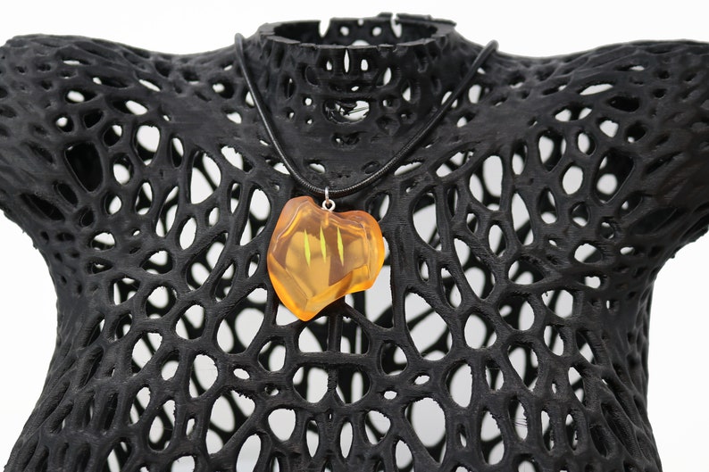 May include: A pendant necklace featuring a translucent, amber-colored stone with a unique shape. The stone hangs from a black cord and is displayed on a black, openwork mannequin. The necklace is a piece of jewelry.