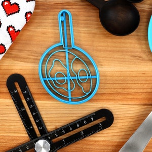 May include: A collection of kitchen tools on a wooden surface. Includes a blue egg-shaped mold, a black angle ruler, a white oven mitt with red pixelated hearts, a blue spatula, and black measuring spoons. A cooking-themed assortment.