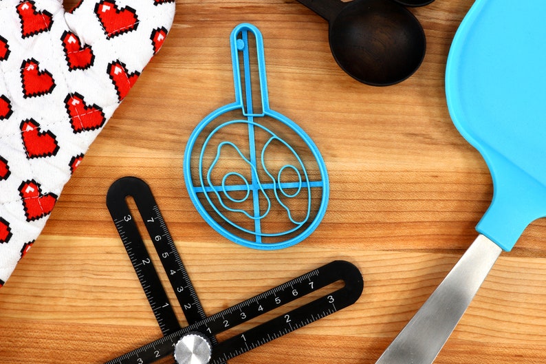 May include: A collection of kitchen tools on a wooden surface. Includes a blue egg-shaped mold, a black angle ruler, a white oven mitt with red pixelated hearts, a blue spatula, and black measuring spoons. A cooking-themed assortment.