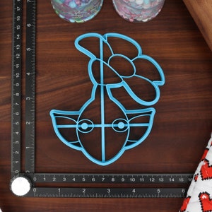 May include: A blue plastic cookie cutter shaped like a cartoon character with a flower on top. The character has large eyes and a wide mouth.