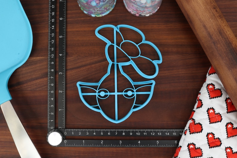 May include: A blue plastic cookie cutter shaped like a cartoon character with a flower on top. The character has large eyes and a wide mouth.