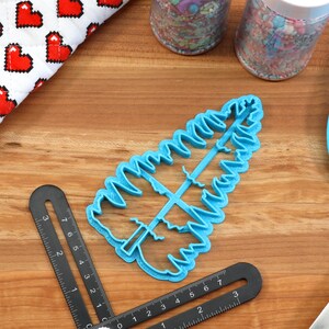 May include: A blue plastic cookie cutter shaped like a Christmas tree. The cutter is on a wooden surface with a ruler and a blue spatula.