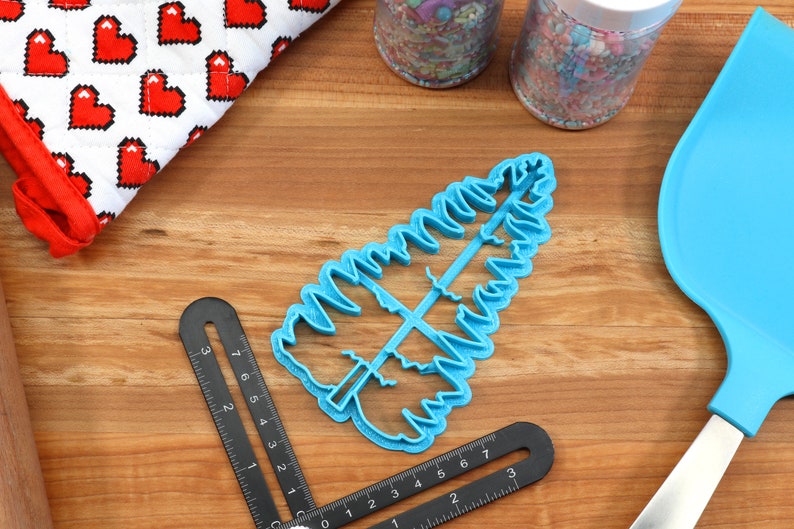 May include: A blue plastic cookie cutter shaped like a Christmas tree. The cutter is on a wooden surface with a ruler and a blue spatula.