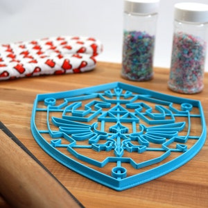 May include: A blue plastic cookie cutter shaped like the Triforce symbol from the Legend of Zelda video game series. The cutter is on a wooden surface with a rolling pin, a red and white kitchen towel, and two jars of sprinkles.