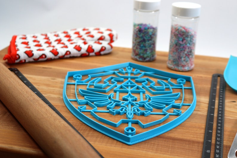 May include: A blue plastic cookie cutter shaped like the Triforce symbol from the Legend of Zelda video game series. The cutter is on a wooden surface with a rolling pin, a red and white kitchen towel, and two jars of sprinkles.