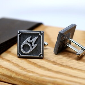K&ouml;nnte beinhalten: Ein Paar silberne Manschettenkn&ouml;pfe mit einem schwarzen und silbernen Feuersymbol auf einem Manschettenknopf. Die Manschettenkn&ouml;pfe befinden sich auf einer Holzoberfl&auml;che.