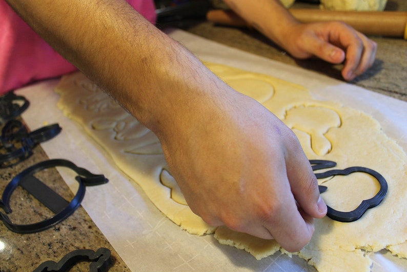 May include: A person using cookie cutters to cut out shapes from a sheet of dough on a white surface.