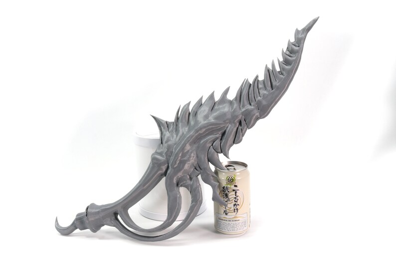 May include: A gray 3D printed sword with a sharp, jagged blade and a curved handle. The sword is resting on a white surface next to a can of Japanese beer.