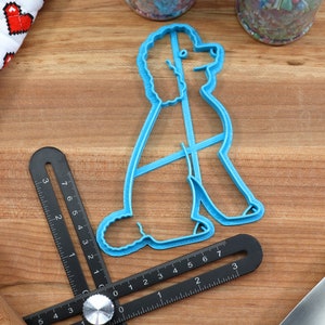 May include: A blue plastic cookie cutter shaped like a poodle sitting down. The cutter is on a wooden surface with a ruler and a rolling pin.