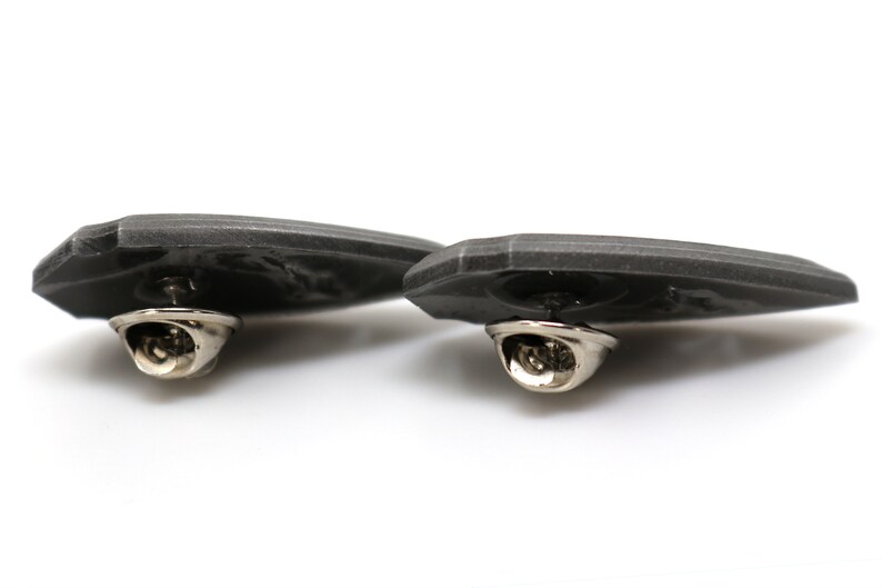 May include: Two gray metal pins with a detailed design. The pins have a silver-colored clasp on the back.