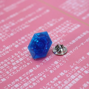 May include: A blue plastic pin with a hexagonal shape and a silver pin back. The pin is on a pink surface with Japanese text.
