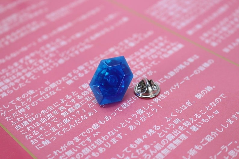 May include: A blue plastic pin with a hexagonal shape and a silver pin back. The pin is on a pink surface with Japanese text.