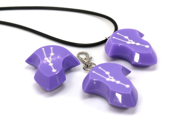 May include: Three purple pendants with a white constellation design. The pendants are shaped like a teardrop and have a shiny finish. One pendant is attached to a black cord necklace, while another is attached to a silver key chain.