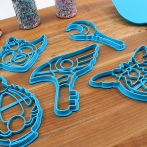 May include: Five blue plastic cookie cutters shaped like different magical wands and staffs. The cutters are on a wooden surface.