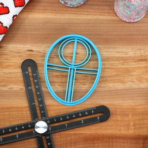 May include: A blue plastic cookie cutter shaped like a circle with a smaller circle inside and a vertical line in the center. The cookie cutter is on a wooden surface.