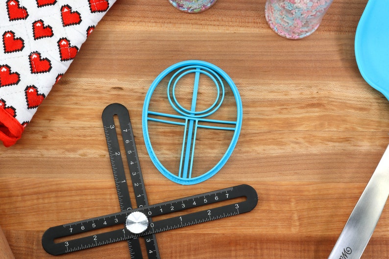 May include: A blue plastic cookie cutter shaped like a circle with a smaller circle inside and a vertical line in the center. The cookie cutter is on a wooden surface.