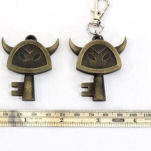 May include: Two gold-colored keychains shaped like keys with a horned helmet design. The keychains are on a white surface with a ruler for scale.