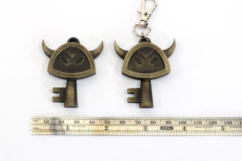 May include: Two gold-colored keychains shaped like keys with a horned helmet design. The keychains are on a white surface with a ruler for scale.