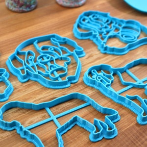 May include: A collection of blue plastic cookie cutters in various shapes, including a top hat, a dog, and a person. The cutters are arranged on a wooden surface, suggesting a baking or crafting theme.