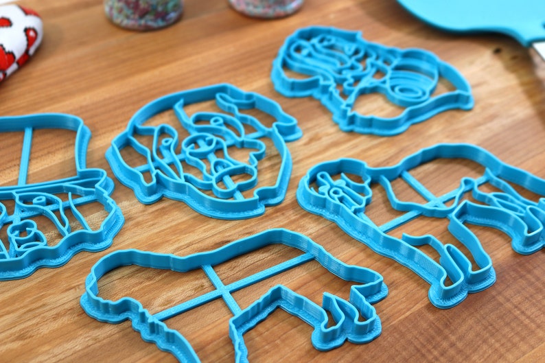 May include: A collection of blue plastic cookie cutters in various shapes, including a top hat, a dog, and a person. The cutters are arranged on a wooden surface, suggesting a baking or crafting theme.