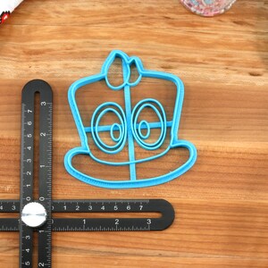 May include: A blue plastic cookie cutter shaped like a cartoon character wearing a hat. The character has two large eyes and a wide smile.