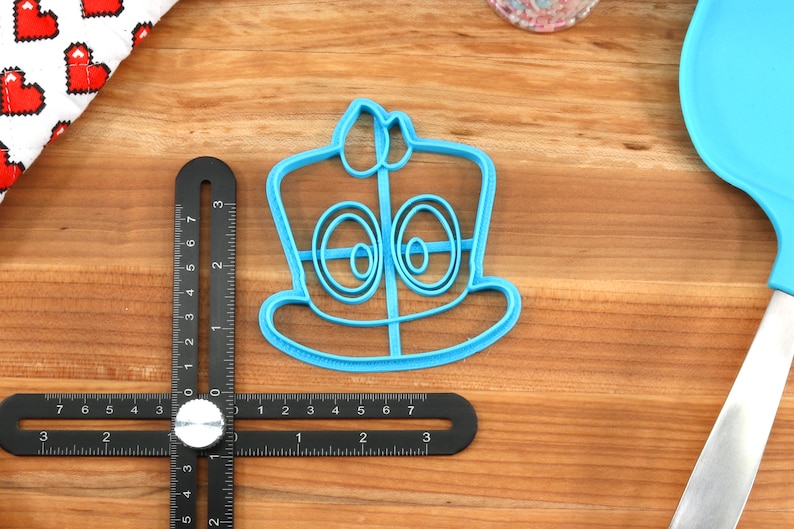 May include: A blue plastic cookie cutter shaped like a cartoon character wearing a hat. The character has two large eyes and a wide smile.